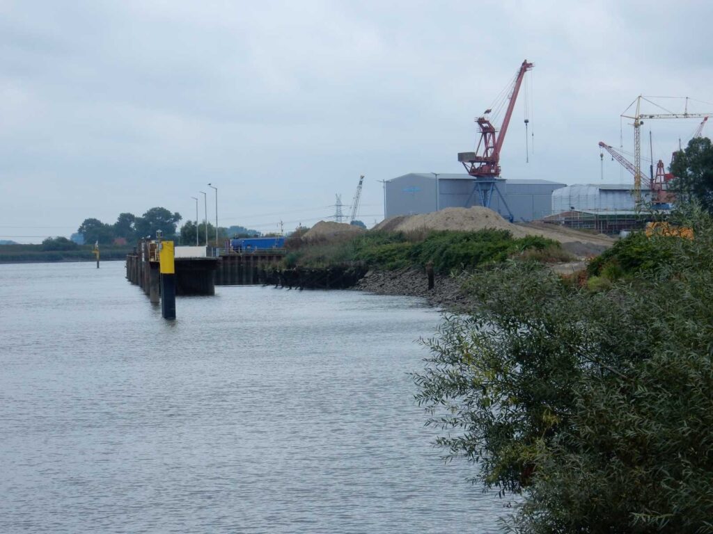 Hafen mit Industrieanlage, Kränen, Sandhaufen und Flussufer – transporttechniker.de