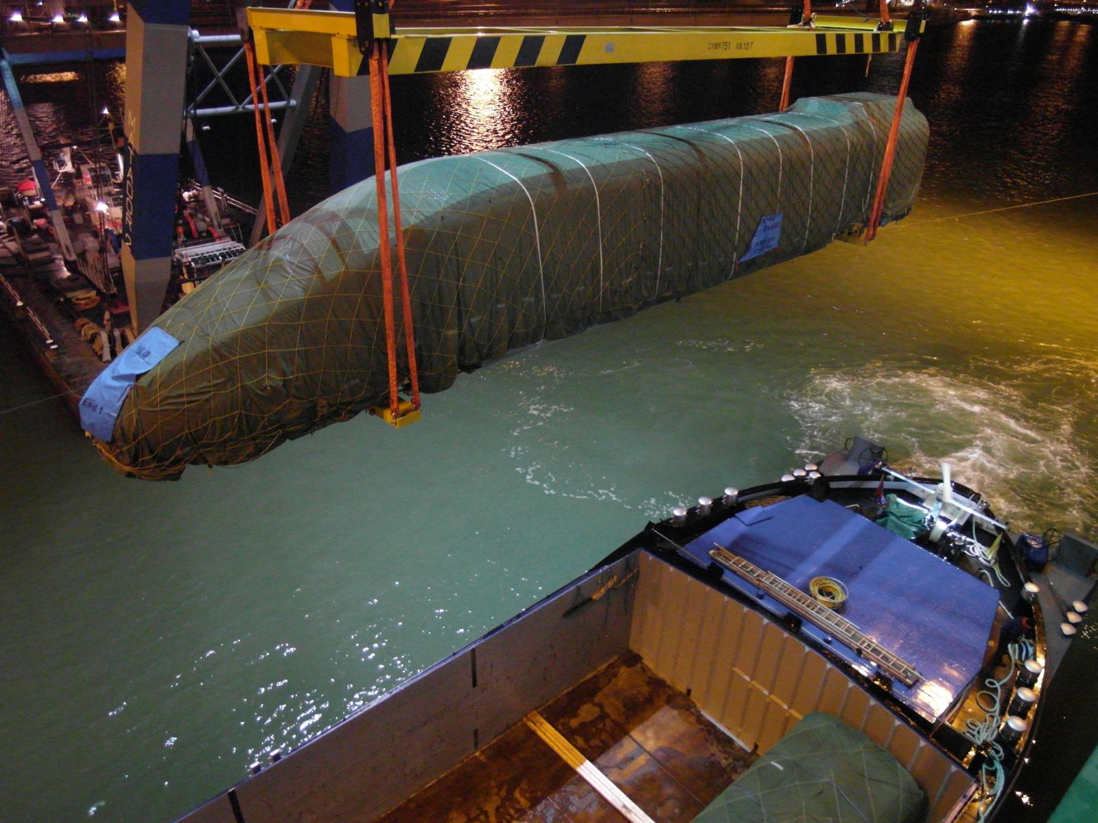 Abgedeckte Zugsektion wird nachts per Schwimmkran über dem Wasser transportiert – grüne Planen schützen die Struktur, im Hintergrund Hafenanlagen mit Beleuchtung und gelb-schwarzer Traverse-transporttechniker-de