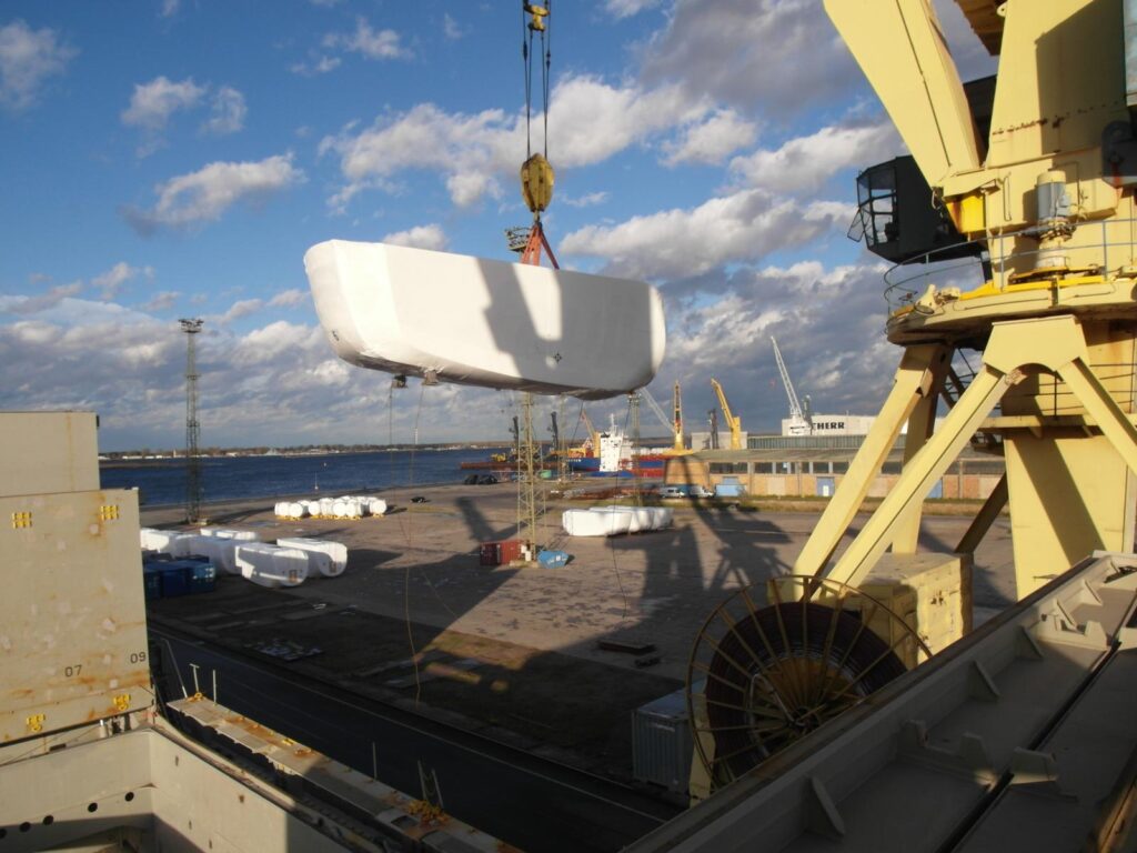 In Folie verpackte Nacelle wird von einem gelben Hafenkran angehoben – im Hintergrund blauer Himmel mit Wolken, Hafeninfrastruktur und weitere Kräne.