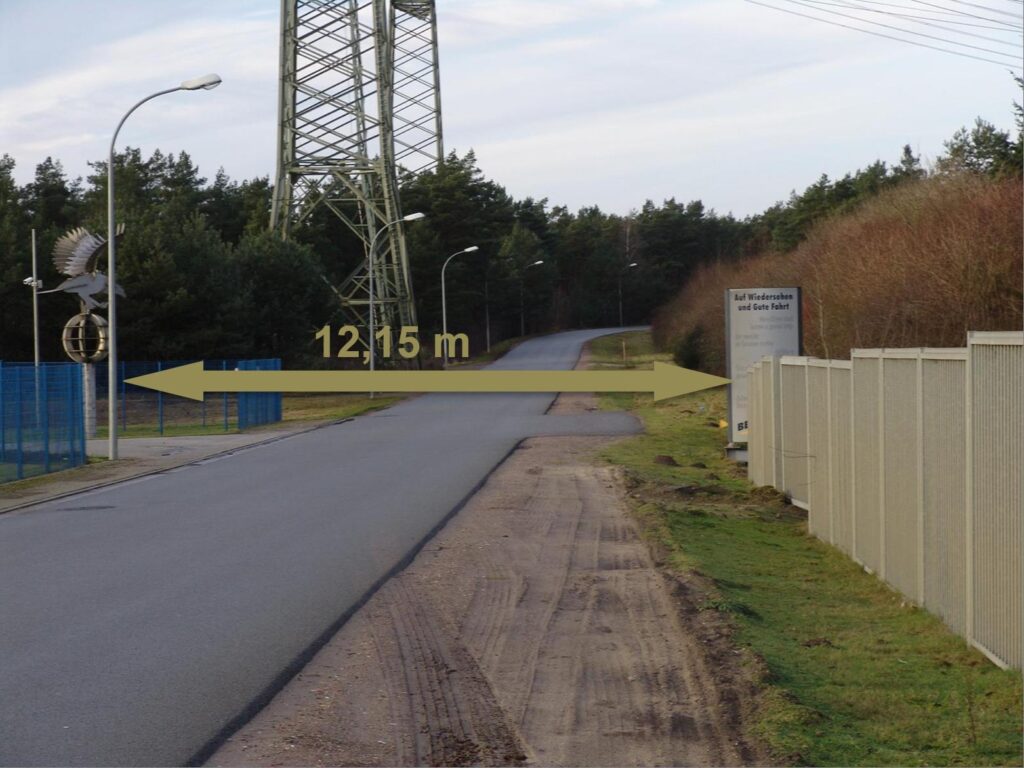 Straßenabschnitt für Schwertransport mit 12,15 m Breite – flankiert von Strommast, Laternen und Waldgebiet, Schilder mit Hinweisen zu Wiedersehen und Guter Fahrt.