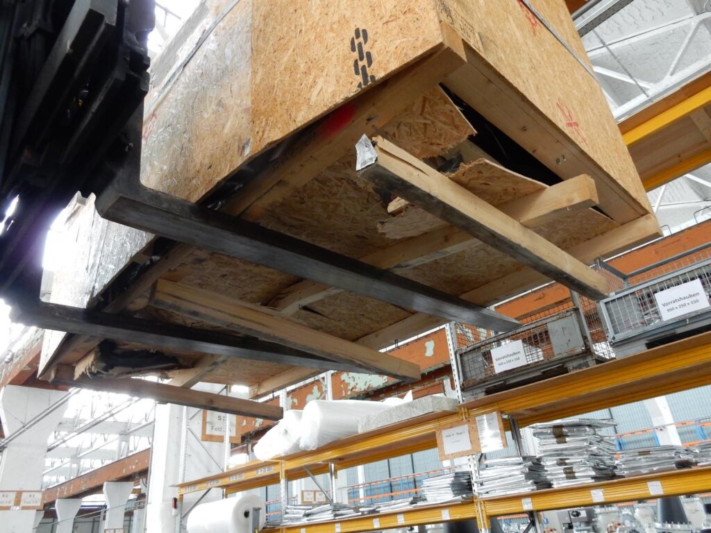 Beschädigte Holzkiste mit gebrochenen Brettern am Lagerregal-Gabelstablerzinken und Hintergrund eines Lagers-transporttechniker-de