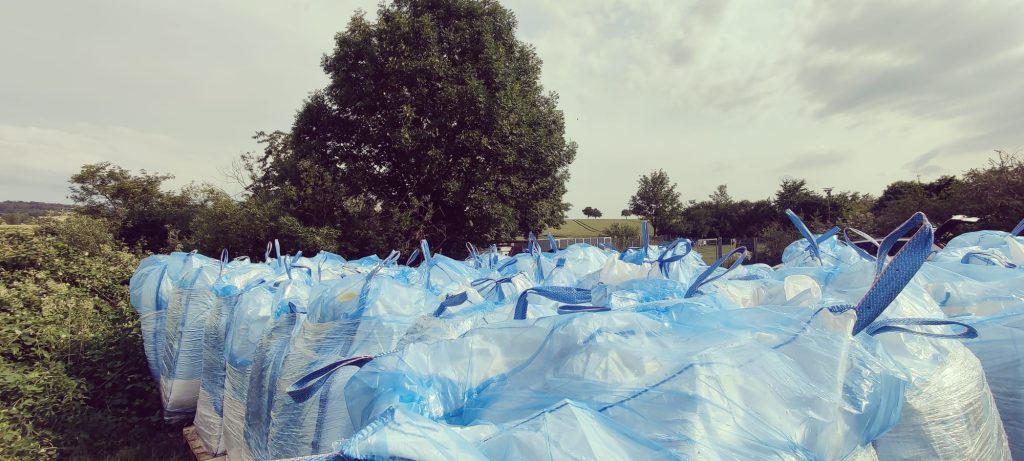 Stapel von blauen Big Bags mit Getreide auf Freifläche – umgeben von Grünflächen und Bäumen unter bewölktem Himmel.