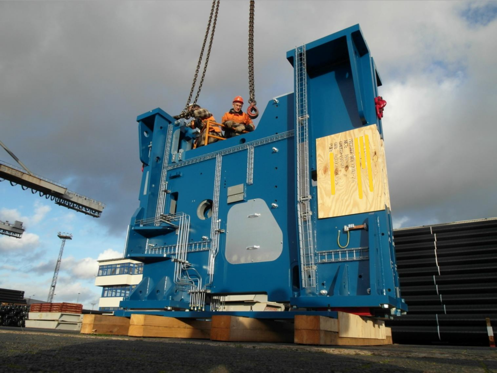 Großer blauer Industrierahmen wird per Kran montiert – Arbeiter in oranger Schutzausrüstung auf der Konstruktion, im Hintergrund Werfthalle und bewölkter Himmel.
