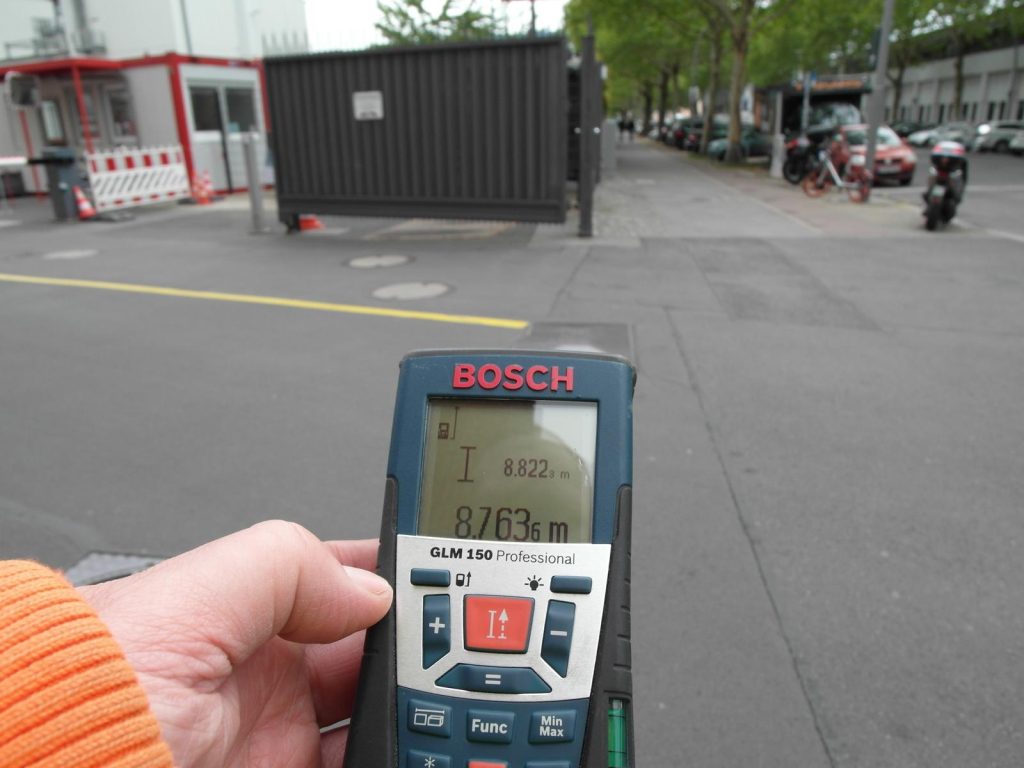 Hand hält Bosch GLM 150 Professional Lasermessgerät zur Abstandsmessung von 8,7636 m an einer Straßenbaustelle mit Containern und Absperrungen