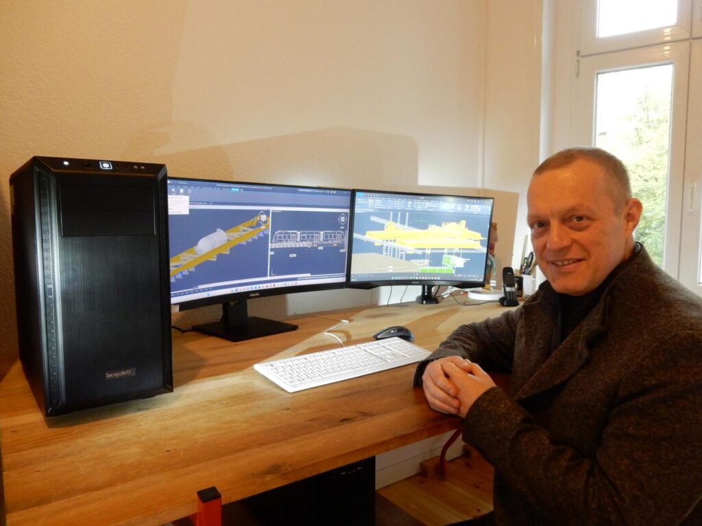 Arbeitsplatz in einem Büro mit Doppelmonitor – 3D-Schiffsbau-Software auf den Bildschirmen, schwarzer PC-Tower (be quiet!), Holztisch, Tastatur und Maus, Mann in grauer Jacke am Schreibtisch