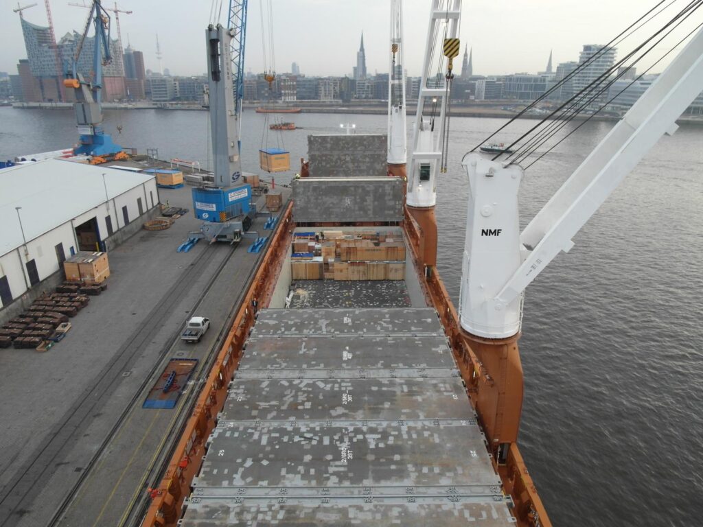 Blick auf Hochseeschiff twindecker während der Beladung im Hamburger Hafen – im Hintergrund Containerkrane, Lagerhallen und die Skyline der Stadt mit Elbphilharmonie