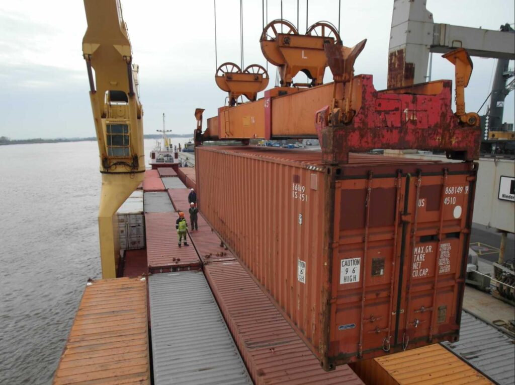 Beladung eines Schiffes mit orangen Containern – Kran hebt Container an, Arbeiter in Schutzausrüstung auf dem Deck, im Hintergrund Wasser und bewölkter Himmel.