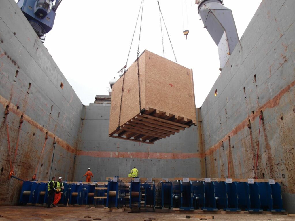 Große Holzkiste wird per Kran in den Schiffsrumpf abgesenkt – Arbeiter in Schutzausrüstung (Helme, Warnwesten) beaufsichtigen den Vorgang.