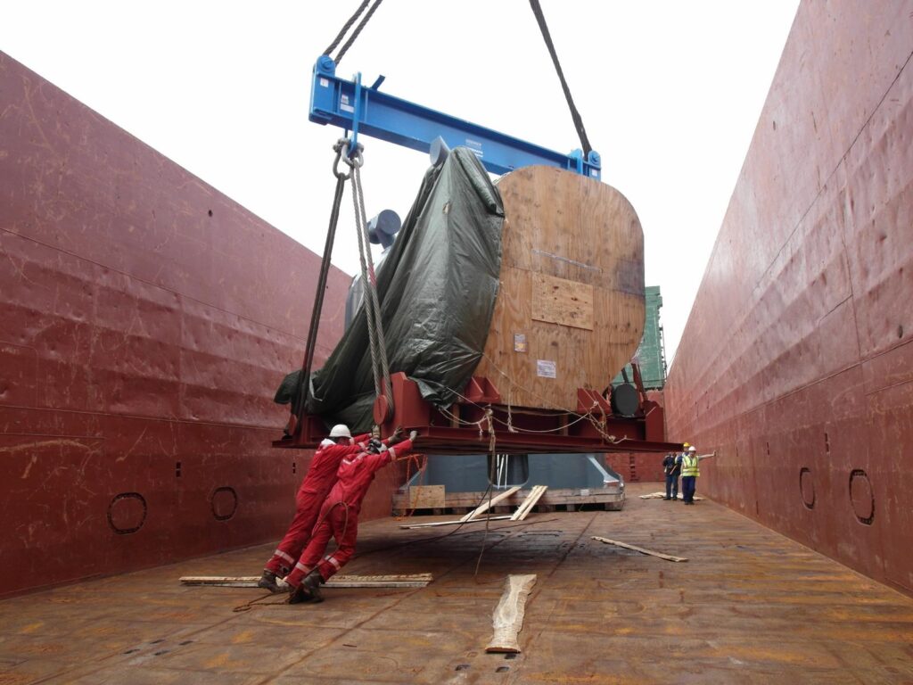 Hafenarbeiter drehen ein Schwerlastteil an einer Traverse im Verladeraum eines Schiffes, um es zu positionieren – im Hintergrund rot-rostige Schiffswand.