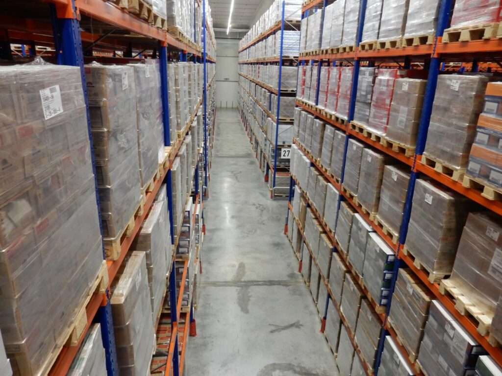 Hochregallager mit beidseitig gestapelten, folienverpackten Kartons auf Holzpaletten – orange-blaue Regalsysteme in einer Industriehalle mit Betonboden.