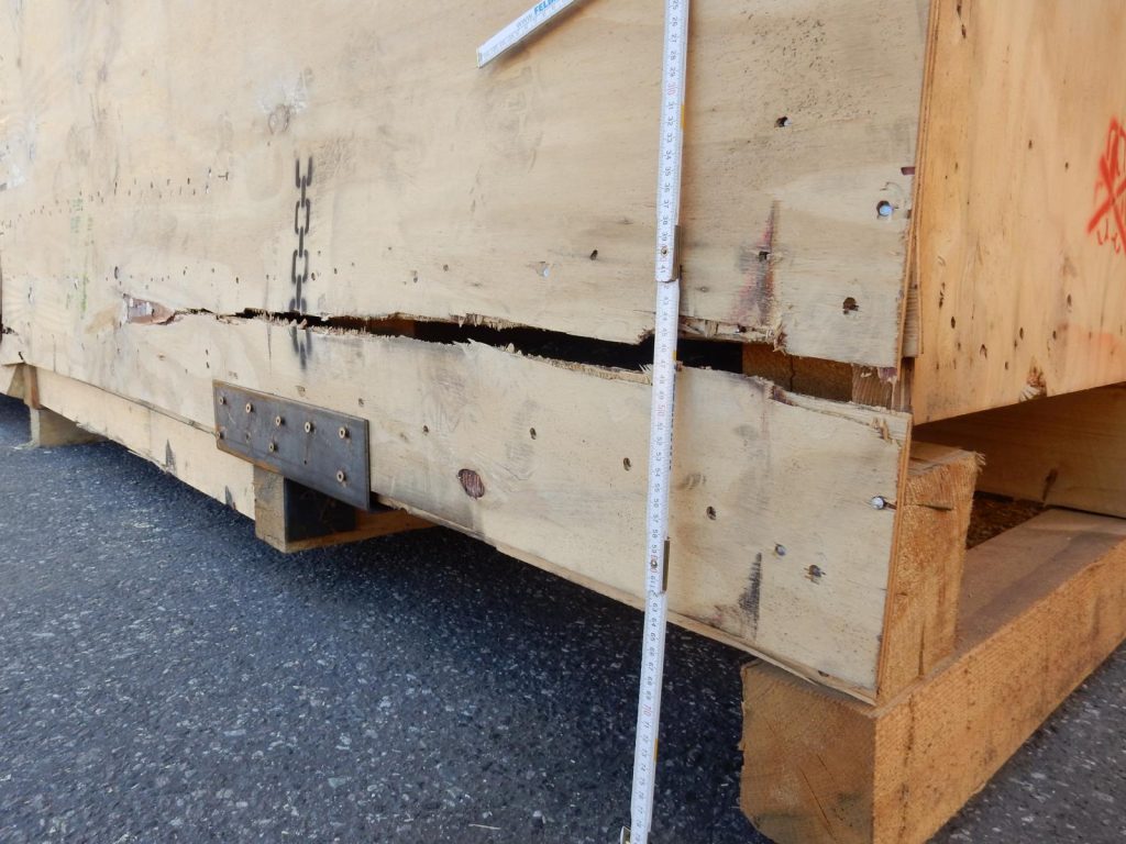 Vermessung einer beschädigten Holz-Kiste mit einem weißen Bandmaß – sichtbare Risse und Absplitterungen an der Kiste, Metallverstärkung an der Unterseite, präzise Ablesung der Maße für Reparatur oder Logistik