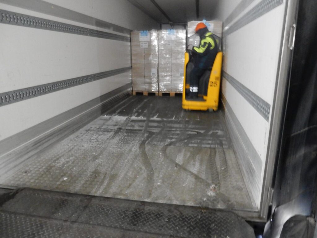 Arbeiter in Warnweste und orangefarbener Mütze bedient einen gelben Gabelstapler im Laderaum eines Kühl-LKWs – folienverpackte Kartons auf Paletten, nasser Boden mit Spuren.