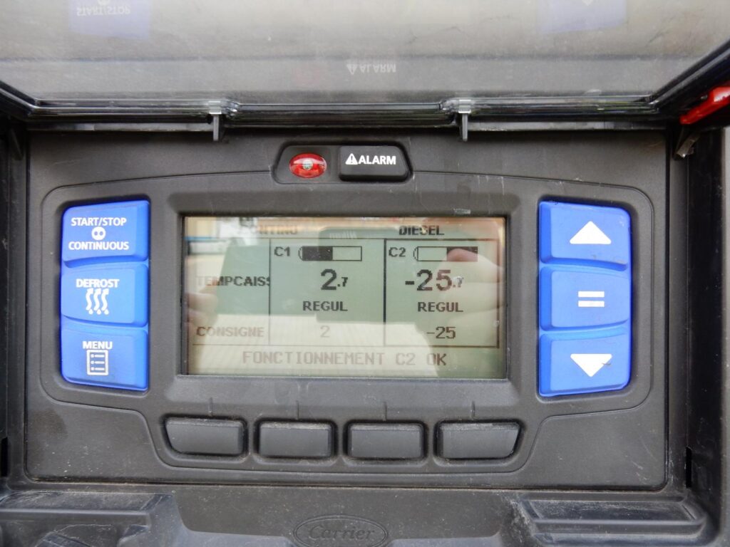Bedienfeld eines Kühlaggregats an einem Kühl-LKW mit blauen Tasten (Start/Stop, Defrost, Menu) – Display zeigt Temperaturen (C1: 2,7°C, C2: -25,7°C) und Statusmeldungen auf Französisch an.