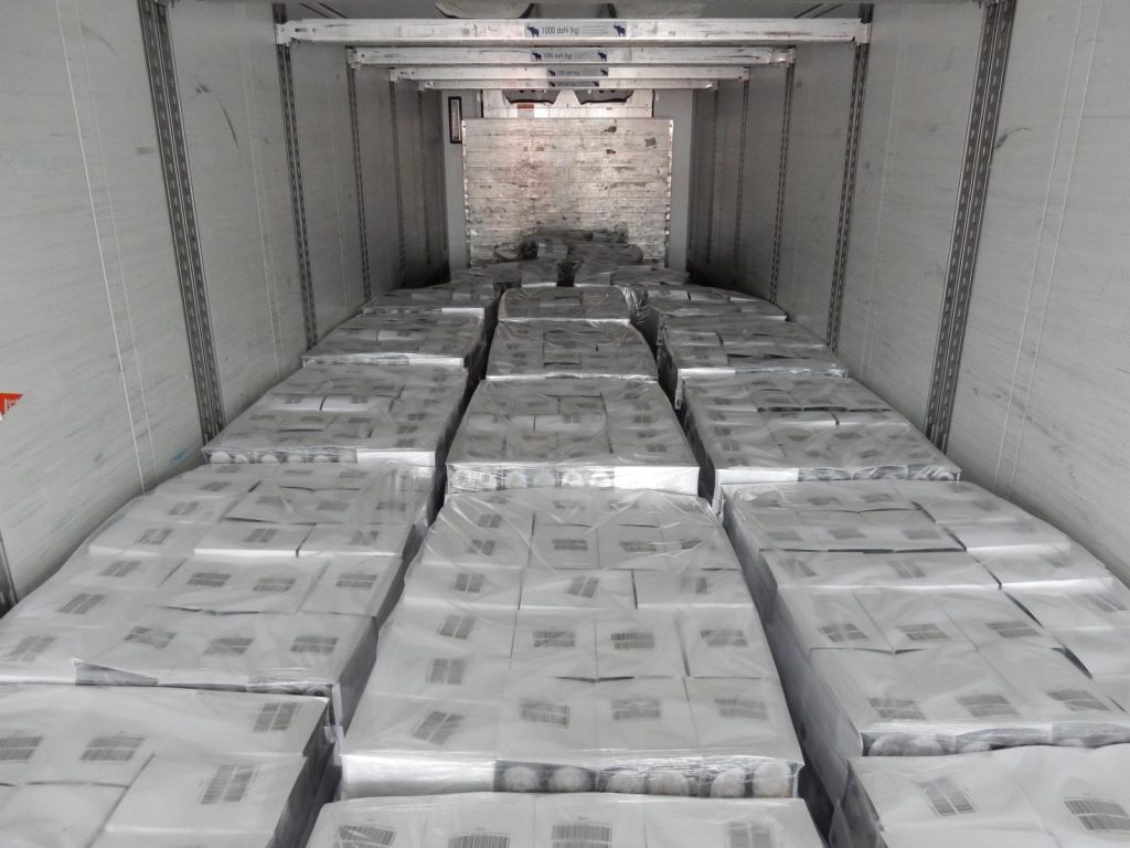 Voll beladener Kühlauflieger mit folienverpackten Kartons auf Paletten – Gewichtsangaben und  nasser Boden und weitere gestapelte Ladung im Hintergrund