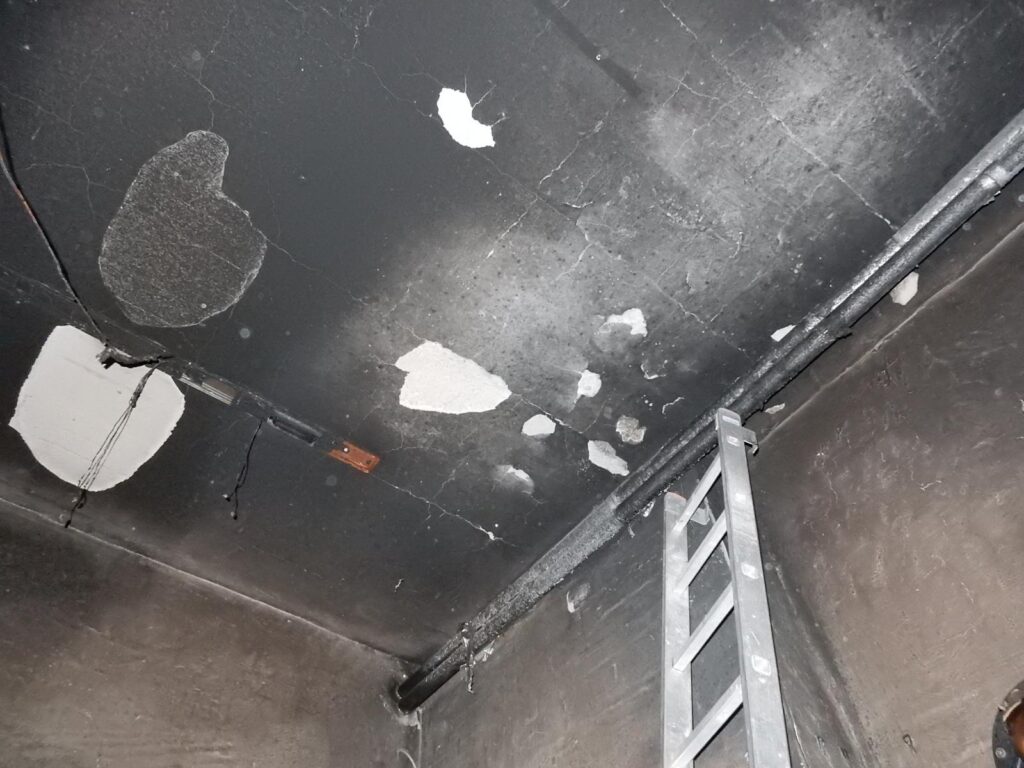 Decke eines Lagerraums nach Brand – stark verrußt mit sichtbaren Abblätterungen und Farbresten, Metallleiter an der Wand, dunkle, verschmutzte Oberfläche