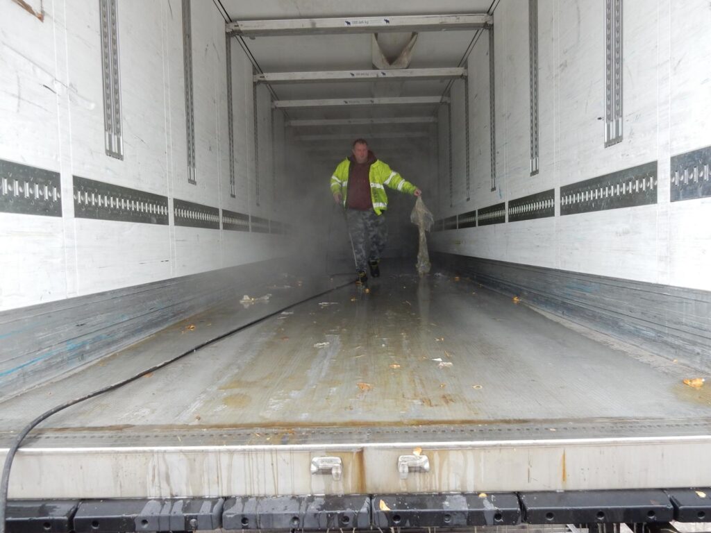 Arbeiter in Warnweste und Schutzjacke reinigt nasse, verschmutzte LKW-Ladefläche mit Besen – offene Ladebordwand im Hintergrund.