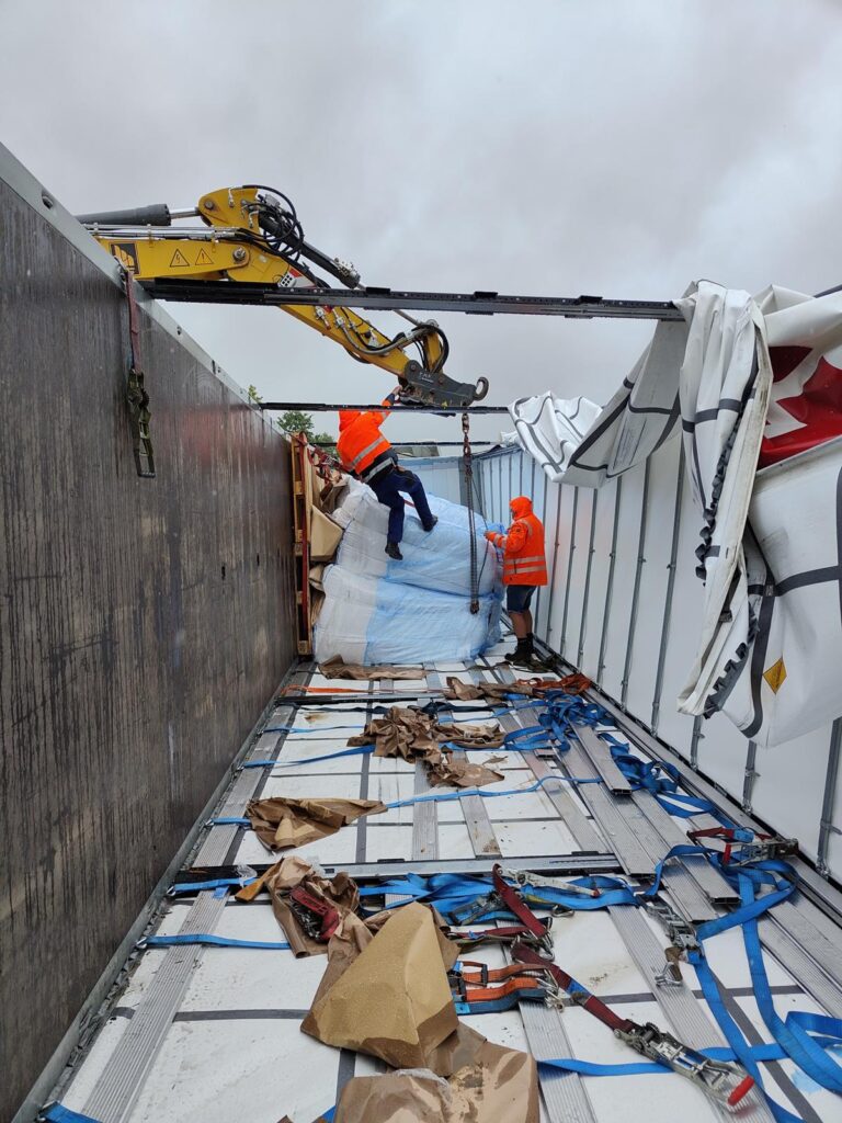 Bergung von verrutschten Big Bags in LKW-Ladefläche – Arbeiter in Warnwesten sichern Ladung mit Kran und Gurten, zerrissene Plane und Spanngurte sichtbar