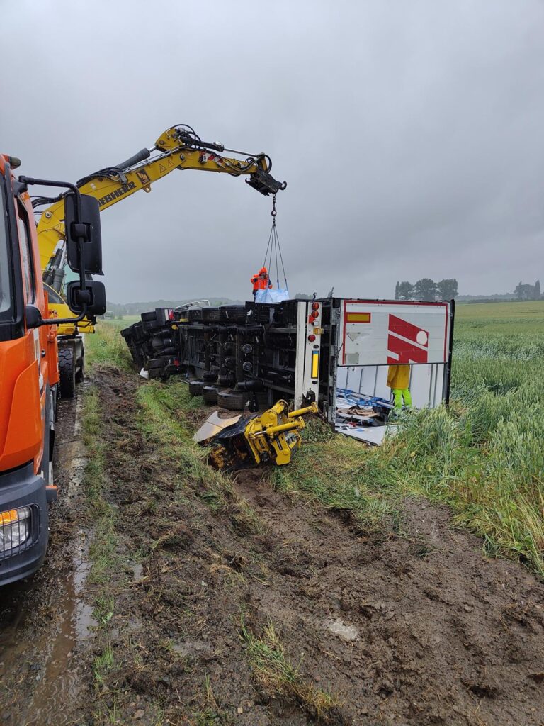 Bergung eines umgestürzten LKW (Schmitz) mit gelbem Kran auf nassem Feld – Arbeiter in Warnwesten heben Teile des Fahrzeugs an, zerrissene Plane und beschädigte Ladefläche sichtbar.