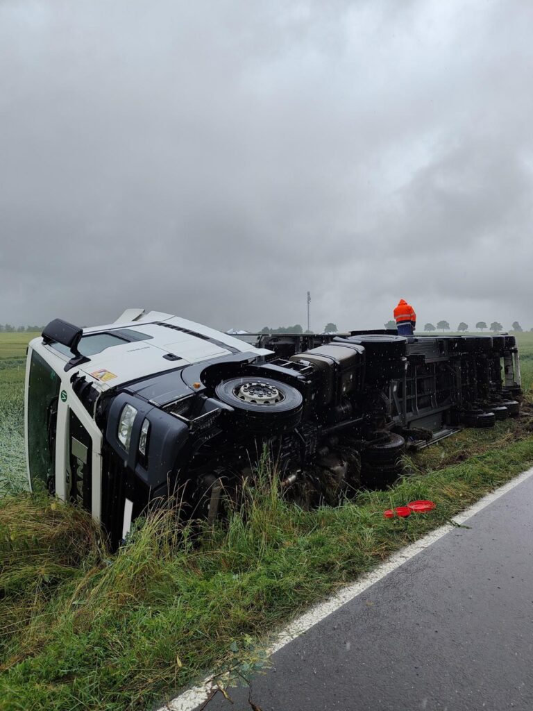 Umgestürzter Sattelzug auf nasser Wiese neben Landstraße bei Regen – Arbeiter in Warnweste steht auf der Ladefläche, Fahrerkabine stark beschädigt.