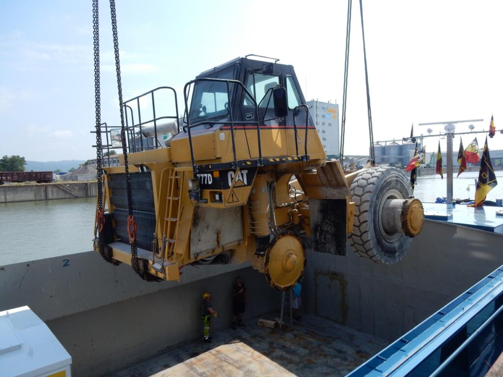 Muldenkipper von Caterpillar wird auf ein Schiff verladen – Hafen mit Flaggen und Industriegebäuden im Hintergrund – transporttechniker.de