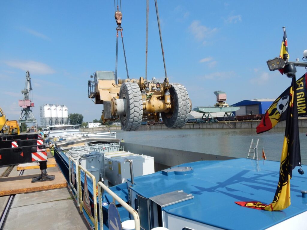 Muldenkipper wird mit Kran auf ein Schiff verladen – Hafen mit Industrieanlagen und Flaggen im Hintergrund – transporttechniker.de