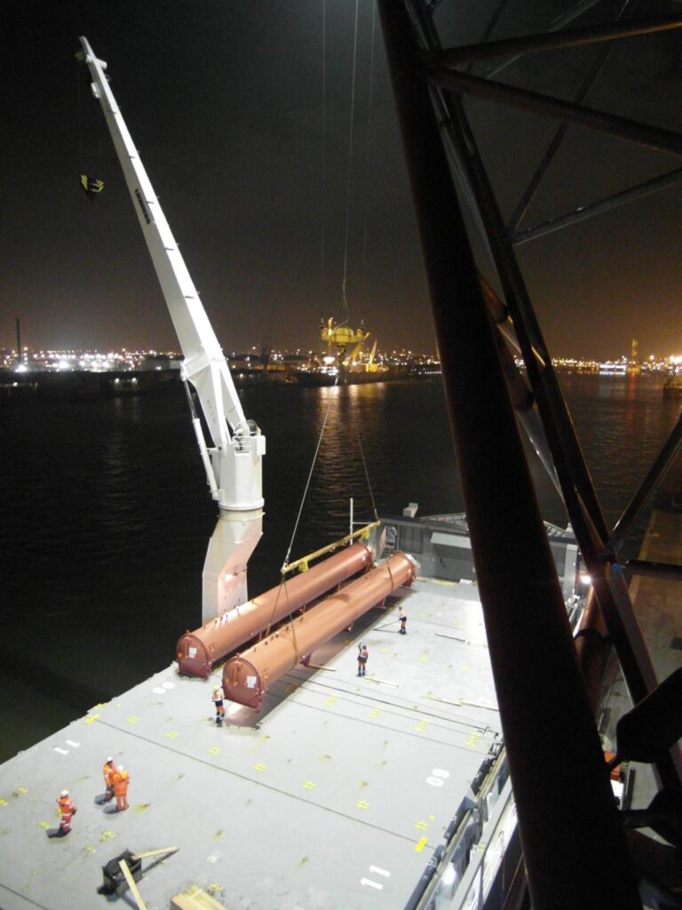 Nachtaufnahme: Kran belädt lange, zylindrische Rohre auf ein Schiff im Hafen – im Hintergrund beleuchtete Stadt und Industrieanlagen am Wasser.