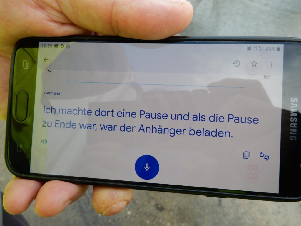 Hand hält Samsung-Smartphone mit geöffneter Germanua-App – Text „Ich machte dort eine Pause und als die Pause zu Ende war, war der Anhänger beladen.“ wird angezeigt, Mikrofon-Symbol aktiv.