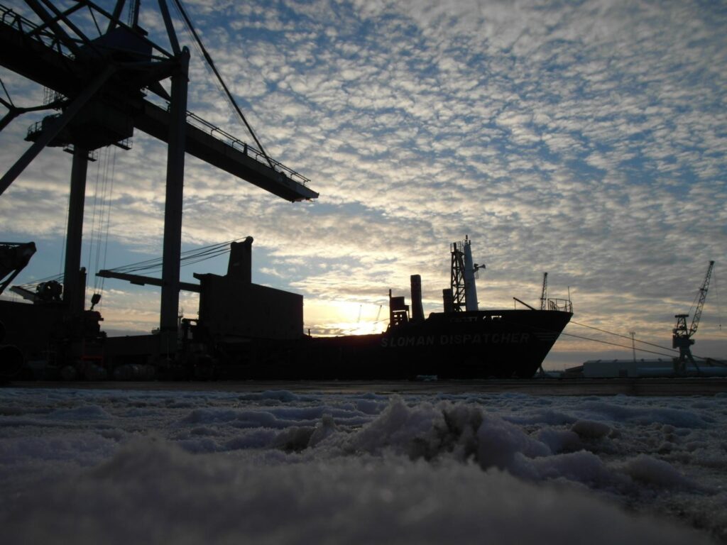 Silhouette des Schiffes "Sloman Dispatcher" bei Sonnenuntergang im verschneiten Hafen – im Hintergrund Containerkrane und bewölkter Himmel.