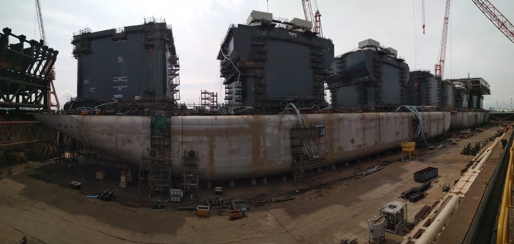 Mehrere Schiffsrümpfe im Trockendock einer Werft – graue Stahlkonstruktionen mit Rostspuren, Gerüste und Kräne im Hintergrund, bewölkter Himmel