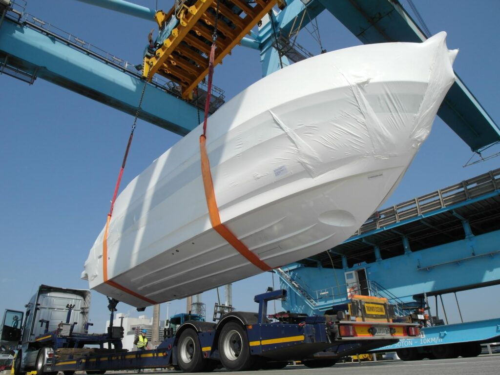 Schiffsbug einer Jacht in Folie verpackt – Entladung vom Lkw-Auflieger per Containerkran mit Traverse auf einen Tieflader, blauer Himmel im Hintergrund.