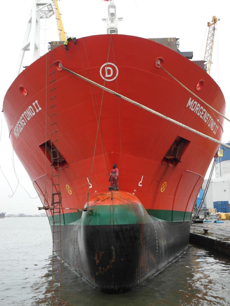 Blick auf den Bug des Schiffes "Morgenstond II" in rot-grün-schwarzer Lackierung – Arbeiter an Seilen und Geländern, im Hintergrund Hafenkran und Wasser.