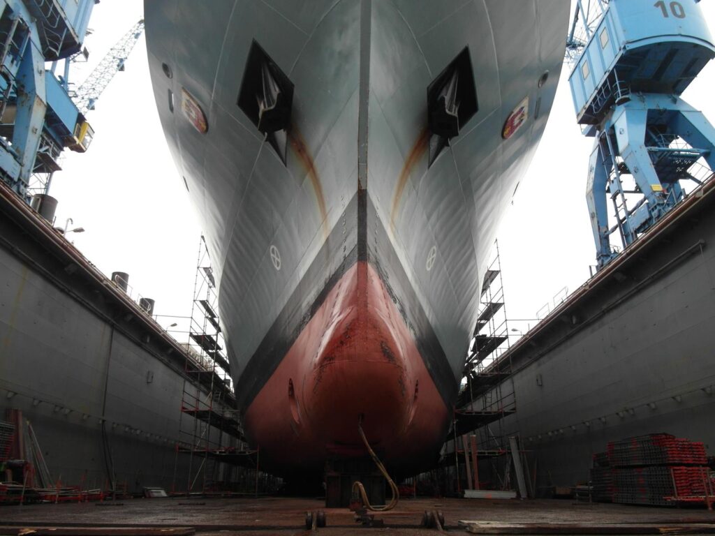 Schiffsbug im Trockendock einer Werft – grauer Rumpf mit rotem Unterwasseranstrich, Rostspuren am Kiel, Gerüste und weitere Schiffe im Hintergrund