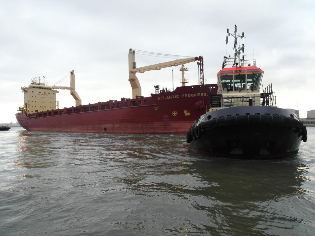 Schwarzes Schleppboot mit roter Brücke begleitet das Frachtschiff Atlantic Progress im Hafenbecken – im Hintergrund Industrieanlagen und bewölkter Himmel