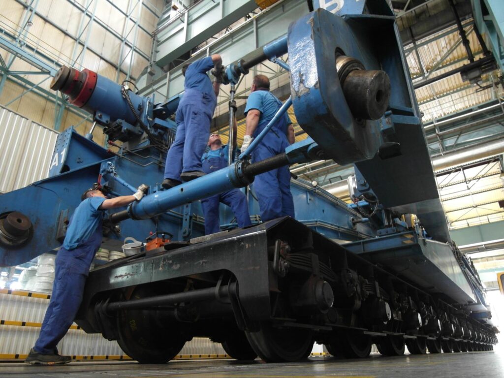Vier Arbeiter in blauer Arbeitskleidung warten einen Schnabelwagen in Industriehalle – Reparatur an Hydraulikzylindern und mechanischen Komponenten.