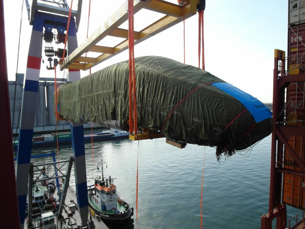 Verpacktes Schnellzugabteil hängt an einer Traverse am Ausleger eines Schwimmkrans ? Verladung in die Containersektion eines Containerschiffs, im Hintergrund Wasser, ein Schubboot und Industriegebäude