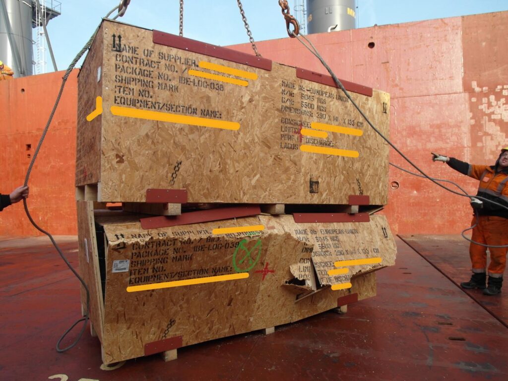 Zwei große Holzversandkisten mit Beschriftungen (u. a. "Made in European Union", "5500 kg", "Dimensions: 290 x 100 x 133 cm") werden per Kran auf dem Schiffdeck transportiert – Arbeiter in oranger Schutzausrüstung leitet den Vorgang.