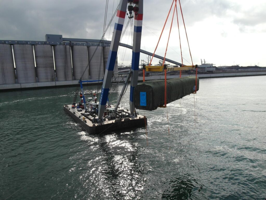 Schwerlastkran auf einem Schiff transportiert eine in grüner Folie verpackte, zylindrische Ladung – im Hintergrund Getreidesilos und Hafeninfrastruktur unter bewölktem Himmel.