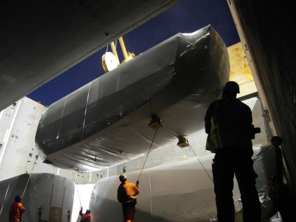 Hafenarbeiter positionieren ein in Folie verpacktes Schwerlastteil per Kran im Verladeraum eines Schiffes – Silhouetten der Arbeiter vor dunklem Hintergrund, weitere verpackte Teile im Raum.