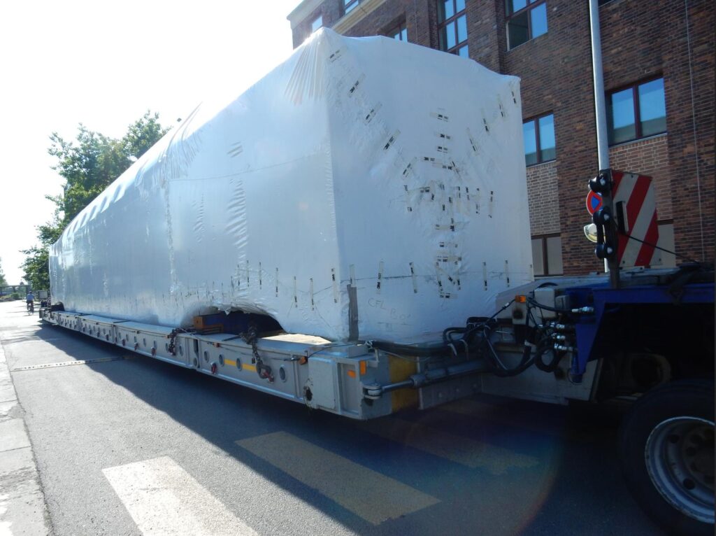 Schwerlasttransport mit in Folie verpackter Ladung auf einem Tieflader – Fahrt durch eine Stadtstraße mit Backsteingebäuden und Verkehrsschildern bei Sonnenschein.