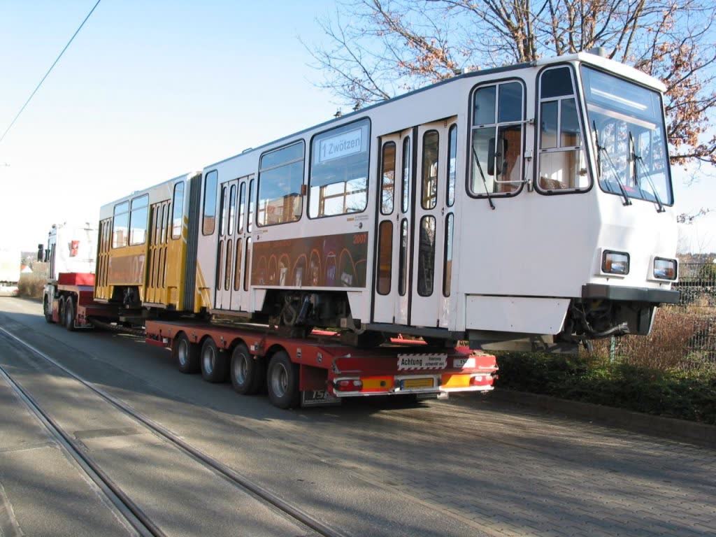 Schwerlasttransport eines Stadtbahn-Waggons auf einem Tieflader mit der Aufschrift "1 Zwötzen" – weißer Waggon mit gelben und roten Elementen, Transport auf einer Straße mit Gleisen, Bäume und blauer Himmel im Hintergrund