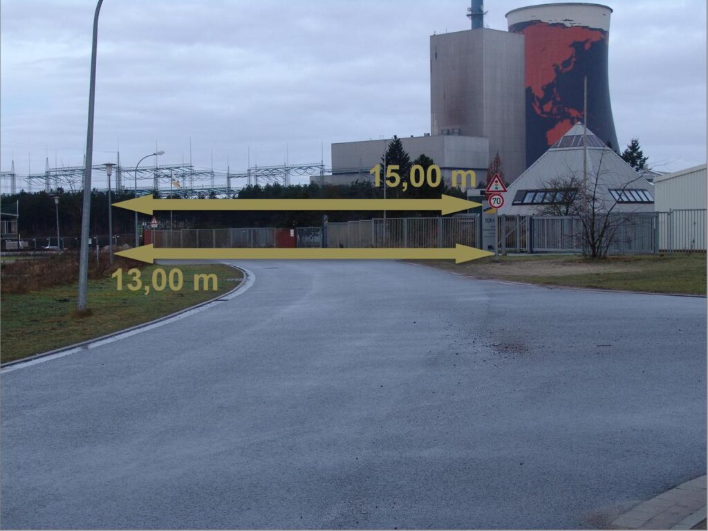 Straßenabschnitt für Schwertransport mit 13,00 m bis 15,00 m Breite vor Industriegebiet und Kraftwerk mit Kühlturm – Verkehrsschilder und Zaunanlage im Hintergrund.