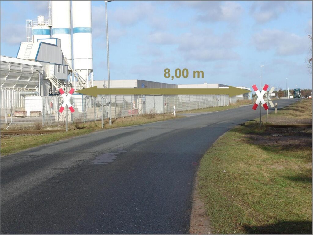 Straßenabschnitt mit 8,00 m Breite für Schwertransport – Bahnübergang mit Andreaskreuzen und Industriegebiet mit Silos im Hintergrund transporttechniker-de