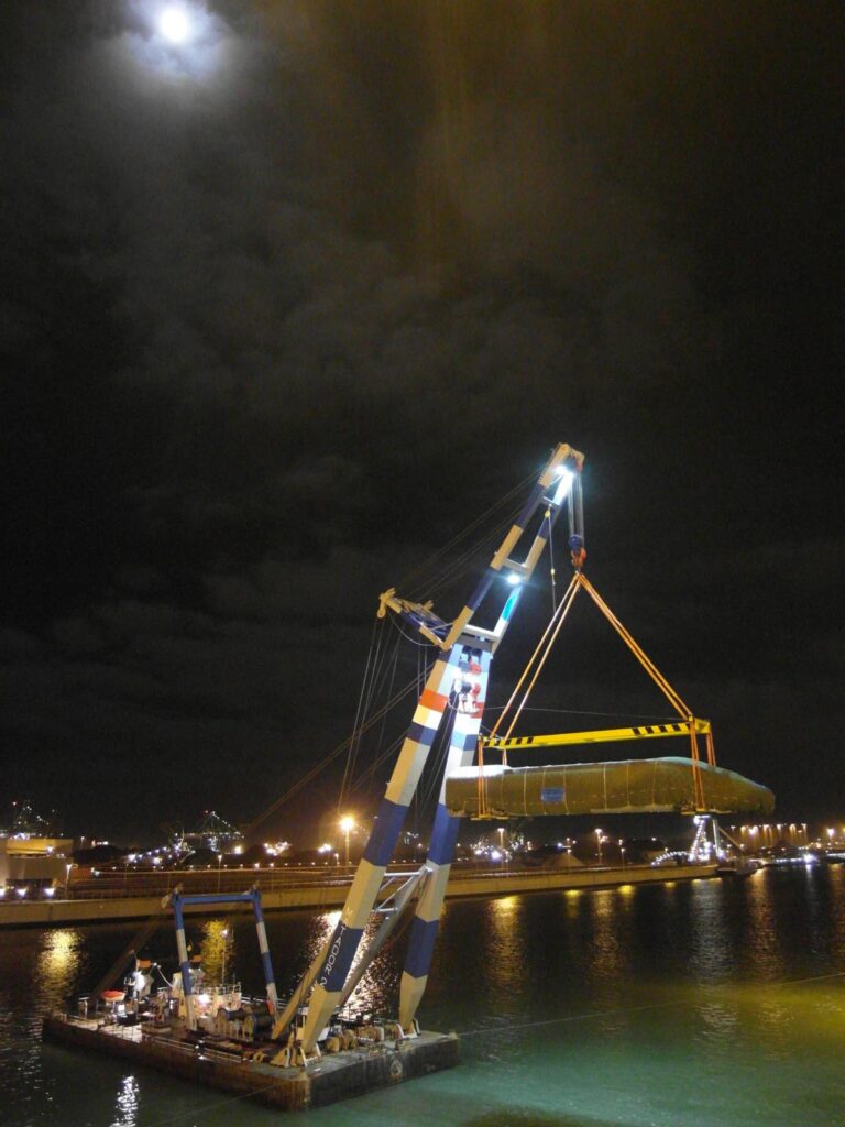 Schwimmkran mit Traverse hebt verpackten Schnellzug als Ladung im Hafenbecken – im Hintergrund Industriegebäude und ein hell erleuchteter Mond am dunklen Nachthimmel-transporttechniker-de