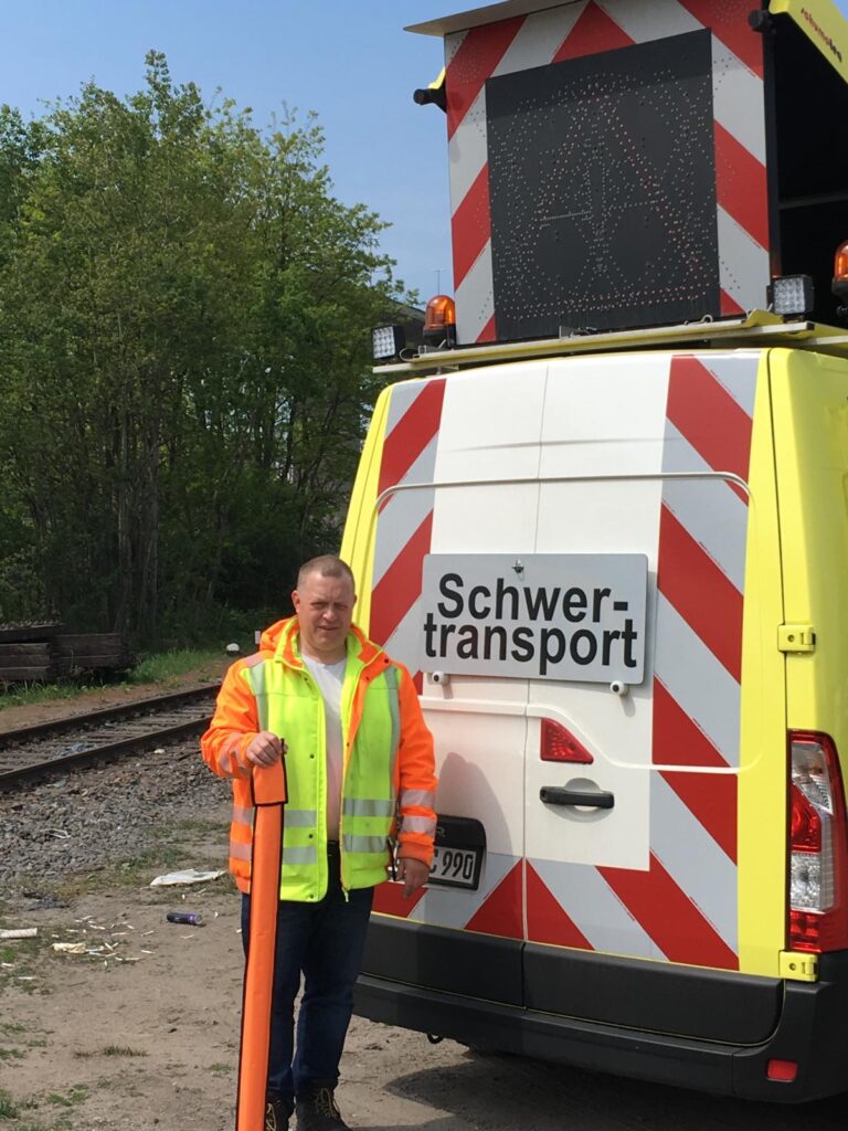 Mann in Warnweste vor Schwertransport-Fahrzeug mit rot-weißen Streifen auf Baustelle mit Gleisen im Hintergrund
