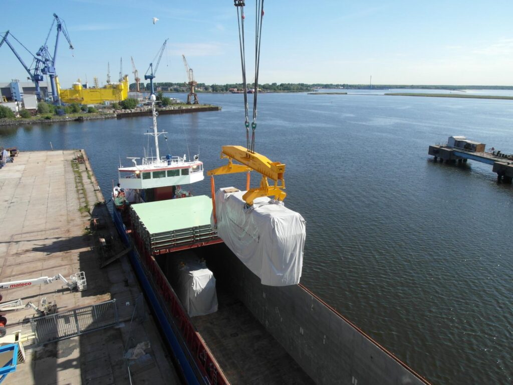 Kran belädt ein großen in folienverpackten  Motor in ein Schiff, im Hintergrund Hafenbecken, Kaianlagen und Containerkrane unter blauem Himmel.