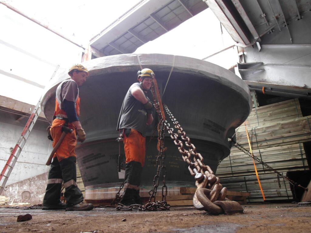 Zwei Hafenarbeiter (Lascher) in oranger Schutzausrüstung und gelben Helmen verzurren einen großen Metallbehälter mit schweren Ketten – im Hintergrund Lower Hold und Zwischendeck eines Schiffes in der Werkshalle.