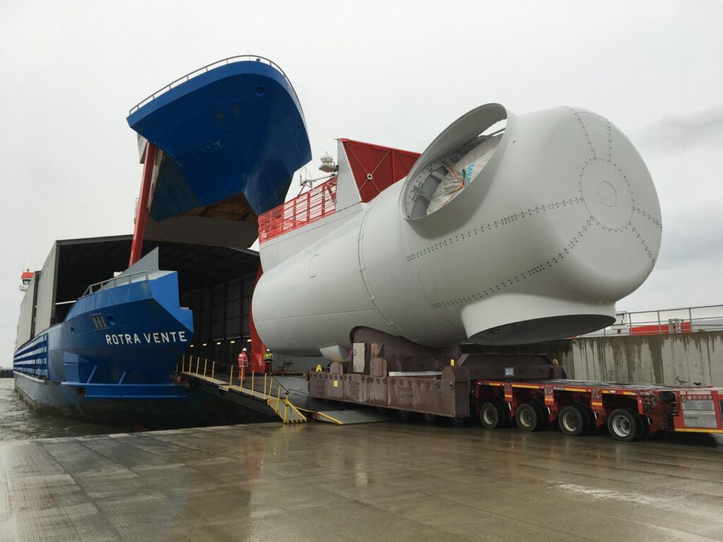 Transport einer riesigen Windkraft-maschienenhaus-nacelle auf einem Tieflader – daneben das Schiff "Rotra Vente" mit geöffneter Laderampe im Hafen.