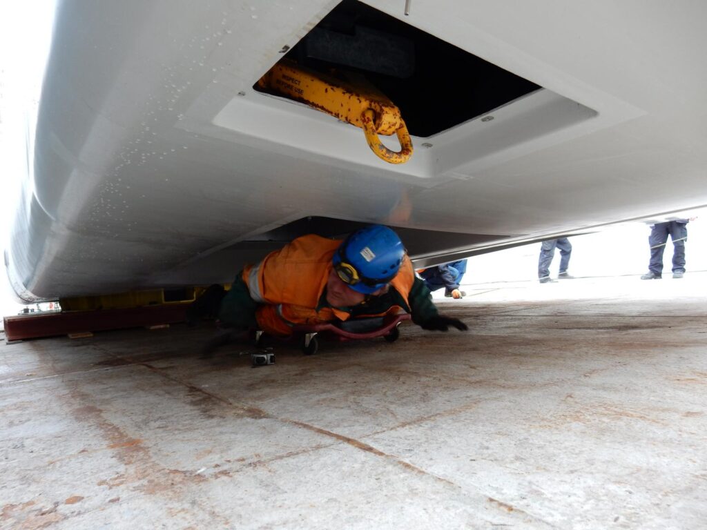 Arbeiter in oranger Schutzausrüstung und blauem Helm inspiziert Nacelle von unten – weitere Arbeiter im Hintergrund, Werftboden mit Markierungen und gelbem Haken an der Schiffswand.