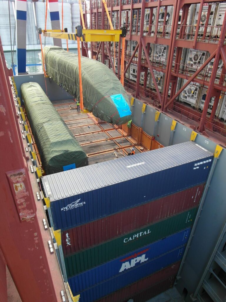 Zugverladung von zwei in grüner Folie verpackten Schiffsaufbauten im Laderaum eines Containerschiffs – Positionierung per Kran, im Hintergrund gestapelte Container.