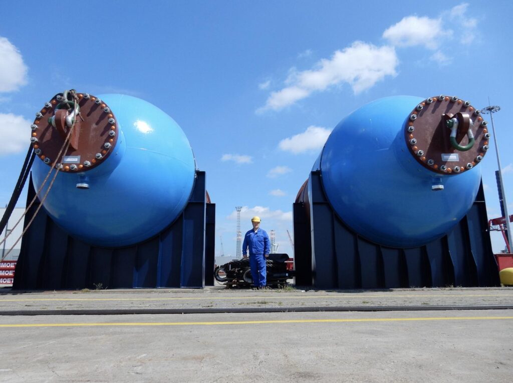 Zwei große blaue Druckbehälter mit rotbraunen Flanschen auf schwarzem Gestell – Arbeiter in blauer Arbeitskleidung und gelbem Helm steht dazwischen, im Hintergrund Hafenkrane und blauer Himmel